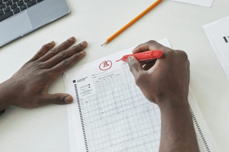 Close-up of a teacher grading multiple choice exams with a red pen on a desk.